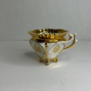 May include: A white porcelain teacup with a gold rim and intricate gold floral designs. The cup has three gold feet and a gold handle.