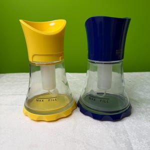 May include: Two clear glass spice grinders with yellow and blue tops. The grinders have a flared base and a curved top. The words "MAX FILL" are printed on the base. The background is a solid green.