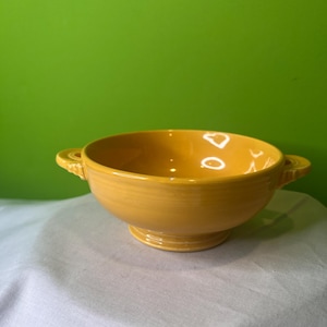 May include: A yellow ceramic bowl with two handles. The bowl is on a white surface.