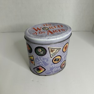Puede incluir: Una lata de metal púrpura con las palabras "The World of the Arts" en la tapa. La lata está decorada con parches coloridos que representan diversas formas de arte, como la música, la danza, el teatro y la fotografía.