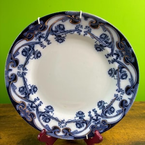 May include: A round ceramic plate featuring a white center and a wide border decorated with dark blue floral patterns and gold-colored accents. The intricate design includes flowing lines and stylized blossoms that frame the plain white basin.