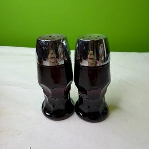 Può includere: Un paio di saliere e pepiere vintage. I contenitori hanno una base in vetro bordeaux scuro e una parte superiore cromata lucida. La base in vetro ha una forma curva unica. Le saliere e le pepiere sono esposte su una superficie bianca con uno sfondo verde.