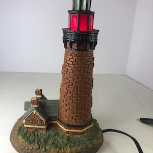 Op de afbeelding: Een rode en zwarte vuurtorenlamp met een bakstenen buitenkant en een klein huis bevestigd aan de basis. De lamp is aan en het licht schijnt door de rode glazen ramen.