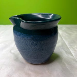 以下が含まれることがあります： 手作りのセラミッククリーマー。濃い緑色の縁と、まだら模様の青色のボディが特徴です。注ぎやすい注ぎ口と丸みを帯びた形状をしています。光沢のある釉薬で仕上げられた陶器です。