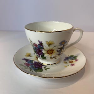 May include: A white teacup and saucer with a floral pattern in shades of purple, yellow, and blue. The cup and saucer have a gold trim.