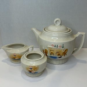 Op de afbeelding: Een vintage set van een witte theepot, roomkan en suikerpot met een grillige illustratie van een schouwscene. De set heeft een crèmekleurige basis met bruine, roze en groene accenten.
