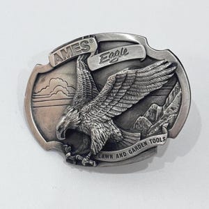 May include: Silver-toned metal belt buckle with an embossed eagle design. The buckle features the text "AMES Eagle LAWN AND GARDEN TOOLS" and a detailed depiction of an eagle in flight over a mountain range.
