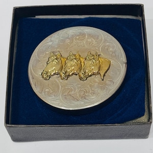 May include: A silver-toned belt buckle with three gold-colored horse heads. The buckle has an engraved floral design and is presented in a black box with a blue velvet lining. Western-themed accessory.