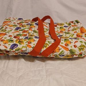 May include: A white tote bag with a colorful vegetable print, featuring carrots, peppers, and eggplants. The bag has bright orange handles and a zipper closure. The bag is designed for carrying groceries or other items.