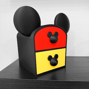 May include: A black storage unit with two drawers featuring a red top drawer and a yellow bottom drawer. The unit has two large black circular ears on top and two smaller black circular knobs shaped like mouse heads on each drawer front.