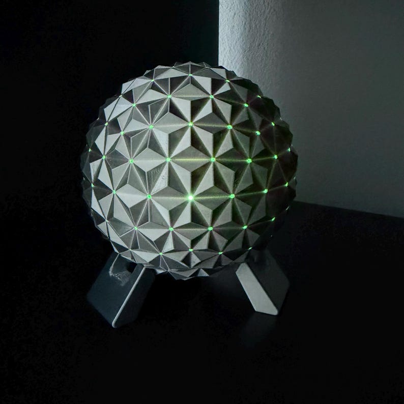 K&ouml;nnte beinhalten: Eine geometrische Kugellampe mit einem grau-wei&szlig;en Dreiecksmuster. Die Lampe hat gr&uuml;ne Lichter und steht auf einer schwarzen Oberfl&auml;che. Die Lampe wird von drei grauen Beinen getragen.