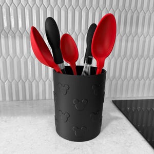 Può includere: Un portautensili da cucina cilindrico nero con un motivo in rilievo di sagome di teste di topo. Il contenitore ospita cinque utensili da cucina assortiti, tra cui cucchiai in silicone rosso, una spatola nera e una piccola frusta in metallo.
