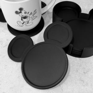 May include: A set of black circular coasters with two smaller circular ears, resembling a mouse head silhouette. One coaster sits under a white ceramic mug featuring a black line drawing of a mouse and the text "all you need is a Smile".