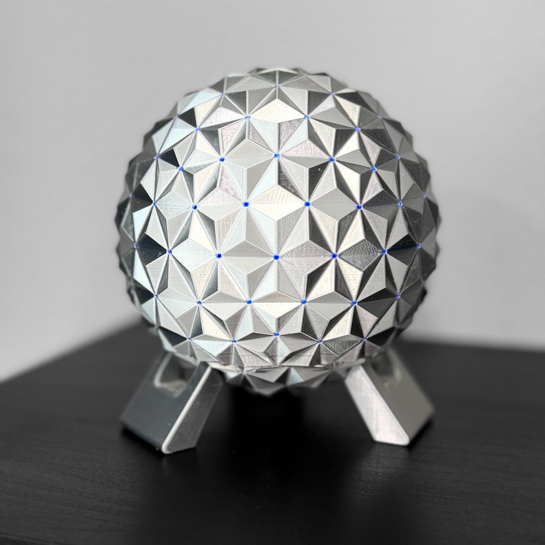 K&ouml;nnte beinhalten: Eine silberne geometrische Kugel mit einem komplexen Dreiecksmuster, die auf einem dreibeinigen St&auml;nder ruht. Die Kugel hat einen metallischen Glanz und kleine blaue Akzente. Das Objekt ist ein dekoratives St&uuml;ck.