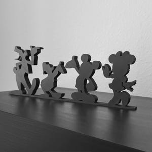 May include: A black silhouette cutout featuring four distinct cartoon character shapes standing in a row on a thin horizontal base. The figures represent a tall character, a duck-like character, a round-eared character, and a bow-wearing character.