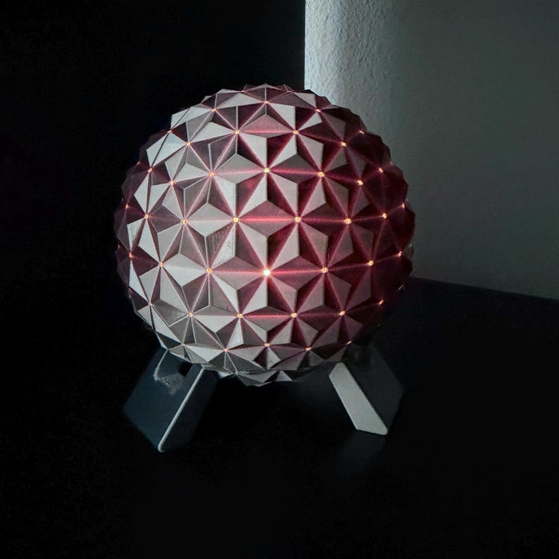 K&ouml;nnte beinhalten: Eine geometrische Kugellampe mit einem komplexen, dreieckigen Muster. Die Lampe ist haupts&auml;chlich grau mit roten Akzenten und steht auf einem dunkelgrauen St&auml;nder. Das Innere leuchtet mit warmem, r&ouml;tlichem Licht und erzeugt einen dekorativen Lichteffekt.