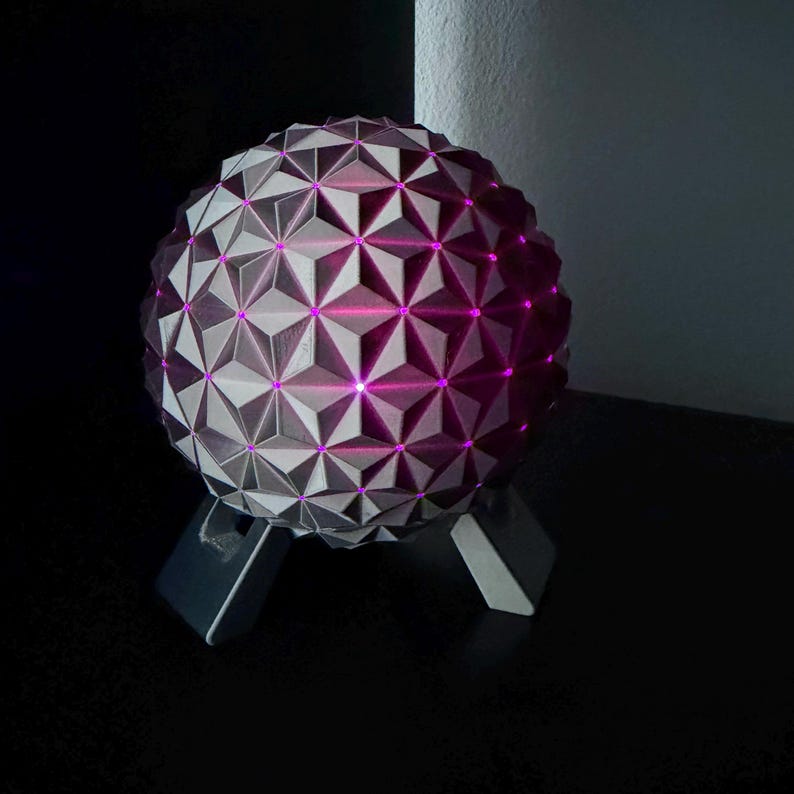 K&ouml;nnte beinhalten: Eine geometrische Kugellampe mit einem komplexen, dreieckigen Muster. Die Lampe ist grau mit rosa Innenbeleuchtung und steht auf einem dunkelgrauen St&auml;nder. Die Lampe steht auf einer schwarzen Oberfl&auml;che.