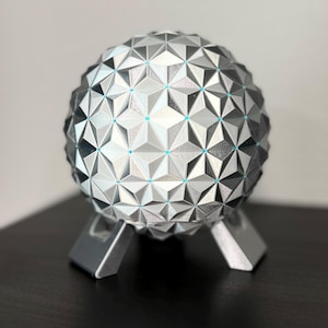 K&ouml;nnte beinhalten: Eine geometrische Kugelskulptur mit einem silbernen und grauen Dreiecksmuster, die auf drei silbernen Beinen ruht. Die Kugel hat kleine blaue Akzente an den Schnittpunkten der Dreiecke. Die Skulptur ist ein dekoratives Objekt.