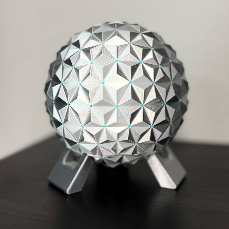 K&ouml;nnte beinhalten: Eine geometrische Kugelskulptur mit einem silbernen und grauen Dreiecksmuster, die auf drei silbernen Beinen ruht. Die Kugel hat kleine blaue Akzente an den Schnittpunkten der Dreiecke. Die Skulptur ist ein dekoratives Objekt.