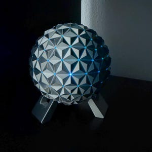 K&ouml;nnte beinhalten: Eine geometrische Kugellampe mit einem grau-wei&szlig;en Dreiecksmuster. Die Lampe hat blaue LED-Leuchten und steht auf einem grauen St&auml;nder. Das Design ist modern und dekorativ.