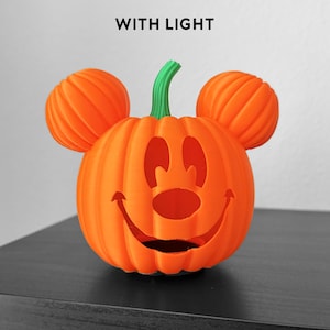Könnte beinhalten: Eine orangefarbene 3D-gedruckte Kürbisdekoration mit grünem Stiel und zwei runden Ohren oben. Die Vorderseite zeigt ein ausgeschnittenes Gesicht, das einer Zeichentrickfigur ähnelt, mit einem breiten Lächeln, ovalen Augen und einer runden Nase.