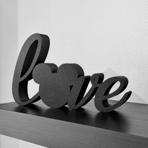 May include: Black, three-dimensional "love" sign with a Mickey Mouse head replacing the "o". The sign is made of a textured material and sits on a dark shelf against a white wall. The letters are in a cursive font.