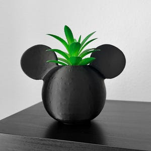 Puede incluir: Una maceta esférica de color negro mate con dos orejas circulares prominentes a los lados, que recuerda a una silueta clásica. El recipiente contiene una planta suculenta artificial de color verde vibrante con hojas puntiagudas y texturizadas.