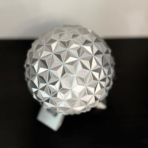 K&ouml;nnte beinhalten: Eine silberne geometrische Kugel mit einem komplexen Muster aus dreieckigen Facetten. Die Kugel hat einen metallischen Glanz und ruht auf einem wei&szlig;en Sockel. Das Design weist kleine blaue Akzente an den Schnittpunkten der Dreiecke auf.