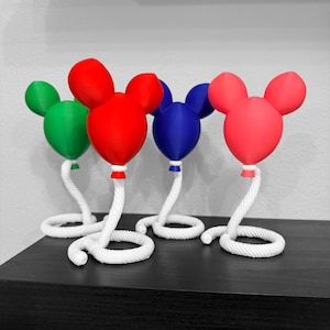 Könnte beinhalten: Vier ballonförmige Skulpturen mit abgerundeten Ohren in verschiedenen Volltonfarben: Grün, Rot, Blau und Pink. Jeder Ballon ist an einem weißen, spiralförmig gewundenen seilartigen Sockel befestigt, der die Struktur aufrecht hält.