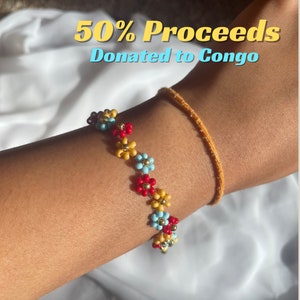 May include: A colorful beaded bracelet with small flower-shaped beads in red, yellow, blue, and white. The bracelet is on a person's wrist. The text "50% Proceeds Donated to Congo" is in the background.