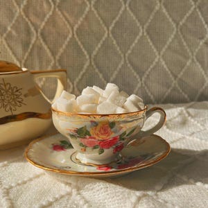 May include: A vintage teacup and saucer set filled with sugar cubes. The teacup features a floral design with gold trim, and sits on a matching saucer. A decorative teapot is visible in the background. The set is placed on a white, textured surface.