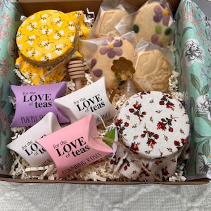 May include: A gift box filled with tea-related items. Includes jars with yellow lids decorated with bees, cookies, tea bags with text "for the LOVE of teas", and a honey dipper. The box has a floral patterned interior.