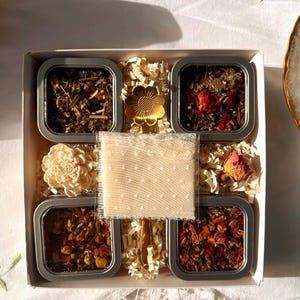 May include: A gift box with four small, square metal tins filled with dried herbs and flowers. The box includes a decorative flower, a small sachet, and shredded paper for cushioning. The contents are arranged on a white surface.