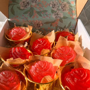 May include: A gift box filled with red rose-shaped soaps in gold-colored holders. The soaps are nestled in brown paper cups and surrounded by shredded paper. The box has a floral patterned interior.