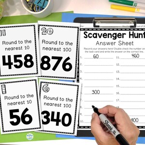 May include: Educational materials for math practice. The image shows task cards with numbers to round to the nearest 10 and 100, along with an answer sheet. A hand is holding a black marker, ready to write answers.
