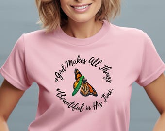 May include: A light pink short-sleeved t-shirt featuring a central graphic of a green caterpillar and an orange and black monarch butterfly. The text "God Makes all Things Beautiful in His Time." is printed in a circular black script around the image.