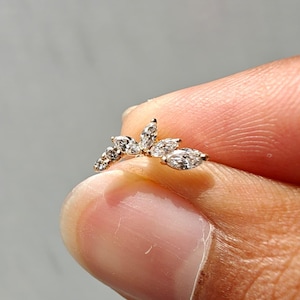 May include: A gold-toned curved stud earring featuring a series of clear, marquise-cut and round-cut stones arranged in a fan-like pattern. The piece is designed with a delicate metal frame that follows the shape of the stones, creating a crown-like appearance.
