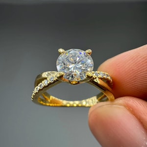 Peut inclure: Une bague en or avec une grande pierre ronde transparente à taille brillant, fixée par quatre griffes. L'anneau présente un design croisé avec de petites pierres transparentes serties pavé le long des deux tiges en or qui se rejoignent.