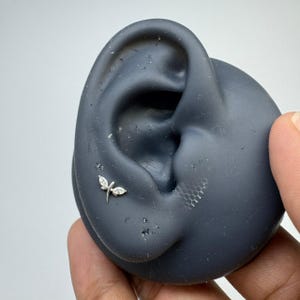 May include: A silver-toned ear stud featuring a dragonfly design with two marquise-cut clear stones forming the wings and a small central stone. The jewelry is positioned on a dark gray silicone ear model to demonstrate its placement and scale.