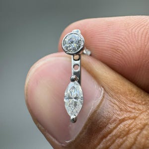 May include: A silver-toned earring jacket featuring a round bezel-set clear stone at the top and a marquise-cut clear stone dangling below. The vertical metal bar connecting the two stones has a small circular cutout detail in the centre.