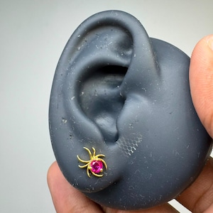 May include: A gold-toned stud earring shaped like a spider with curved legs extending from a central, round, vibrant pink gemstone. The earring is positioned on a dark gray, ear-shaped display form, showcasing the detailed metalwork and stone.