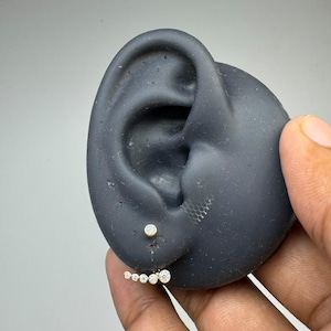 May include: A gold ear climber earring featuring a row of five graduated round clear stones, with a single matching round stone stud positioned above it, displayed on a dark grey silicone ear model to show placement and scale.