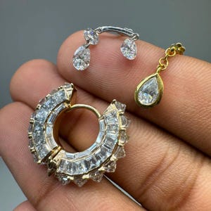 May include: A gold hoop earring encrusted with numerous baguette-cut clear stones, accompanied by two separate dangling charms. One charm is a silver-toned piece with two pear-shaped stones, and the other is a gold-toned piece with a single pear-shaped stone.