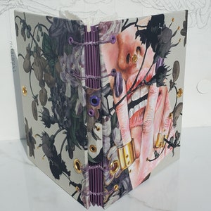 May include: A handmade journal with a floral design and a close-up of a woman's face with a ring on her finger. The journal is bound with purple thread and has gold eyelets.