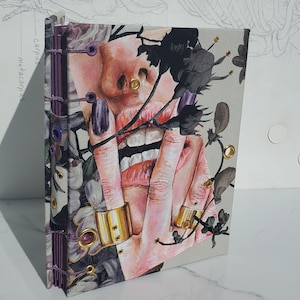 May include: A handmade journal with a floral design and a painting of a woman's face with a ring on her finger. The journal has a purple spine and gold accents.
