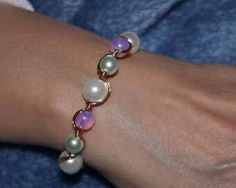 May include: A gold wire bracelet with white, green, and purple beads. The bracelet is on a person's wrist.