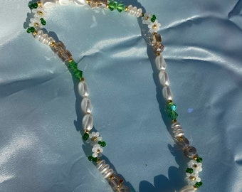 May include: A white pearl necklace with green and gold accents. The necklace features small white flowers and green leaves.