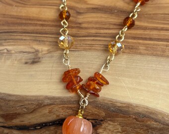 May include: A gold-colored chain necklace featuring a small, orange pumpkin pendant. The necklace is adorned with amber-colored beads and faceted crystals. The necklace is displayed on a wooden surface.