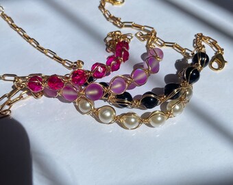 May include: Three gold chain bracelets with different colored beads. The top bracelet has pink beads, the middle bracelet has iridescent purple beads, and the bottom bracelet has white beads.