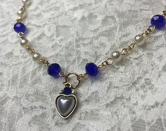 May include: A gold chain necklace with blue and white beads and a silver heart pendant with a pearl center. The necklace is on a white lace background.